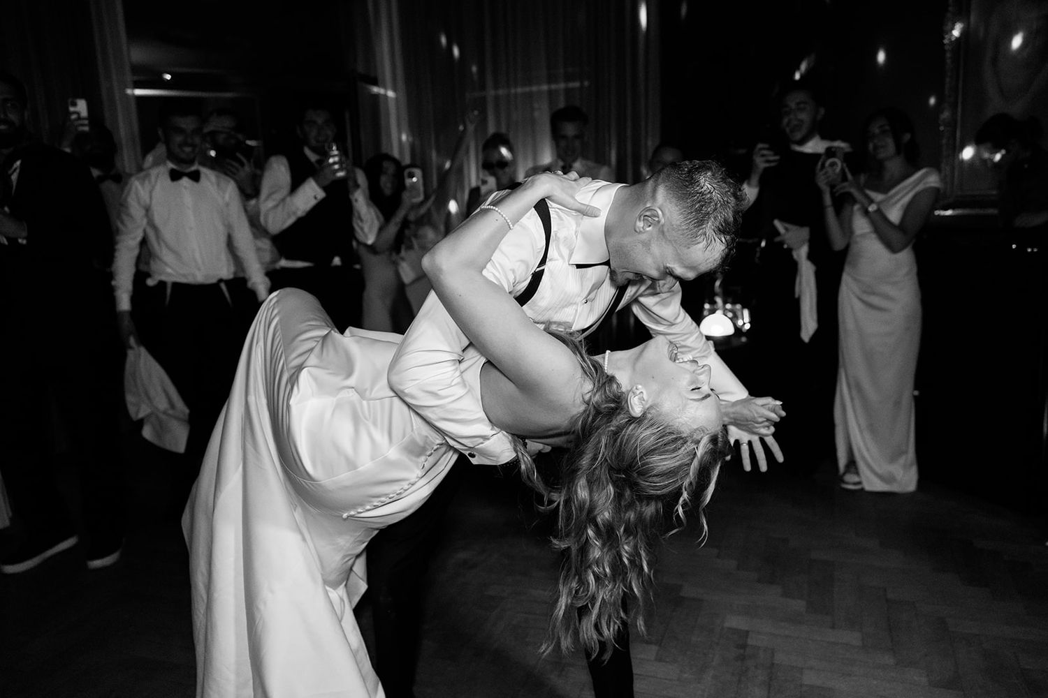 A couple in white wedding attire dances together at their reception as guests watch in the background.