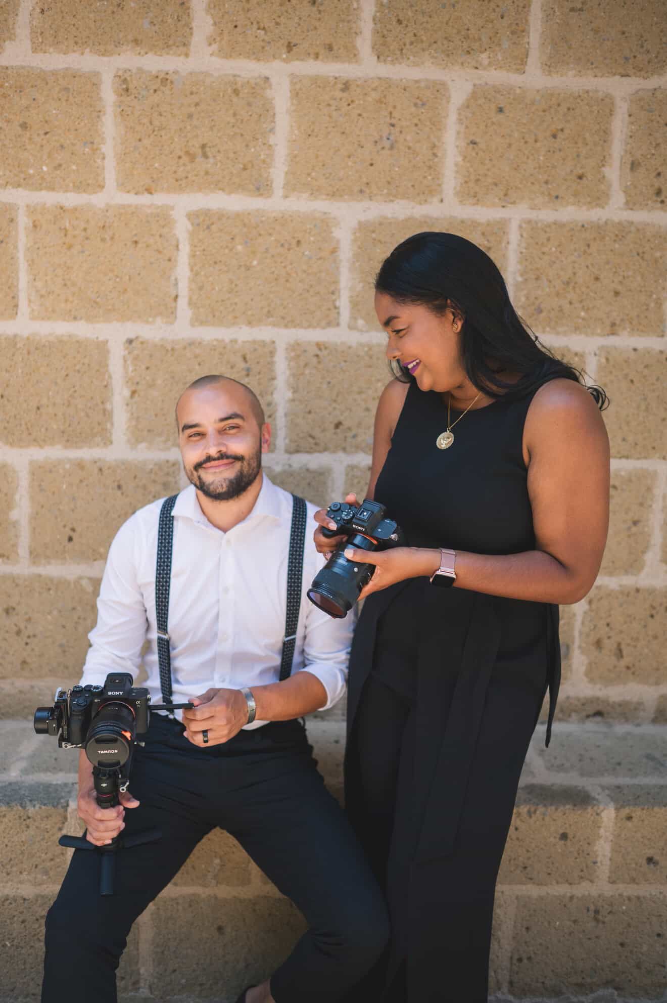 Two photographers in black and white outfits check their cameras while working together against a brick wall.