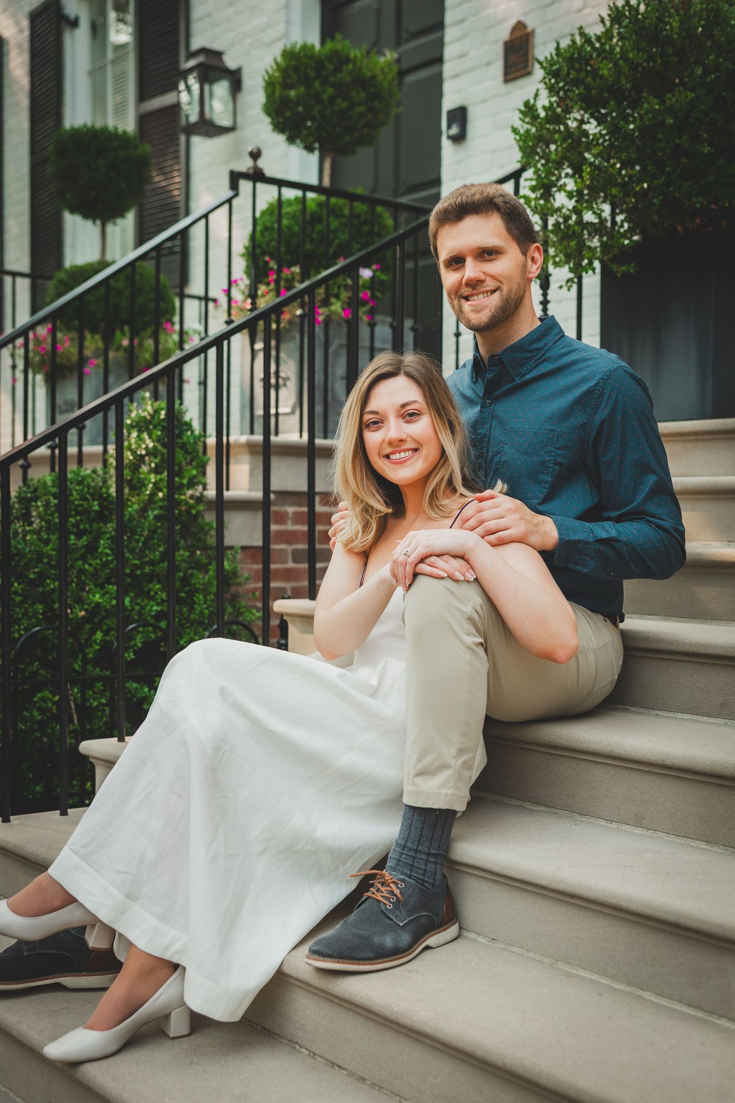 A couple sits together on white stone steps outside a building with decorative topiaries and lanterns.