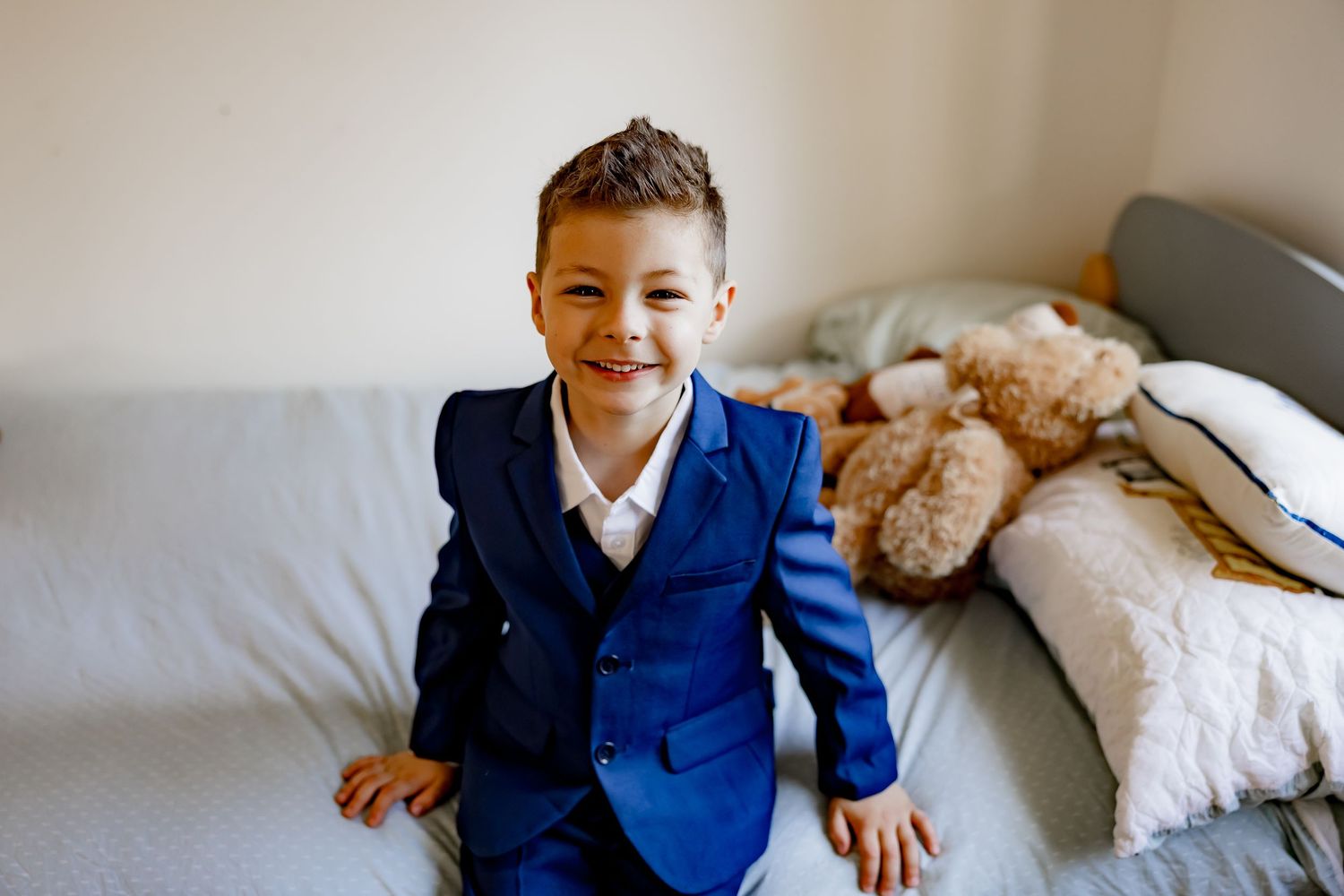 A young child in a navy suit sits on a white bed with decorative pillows.