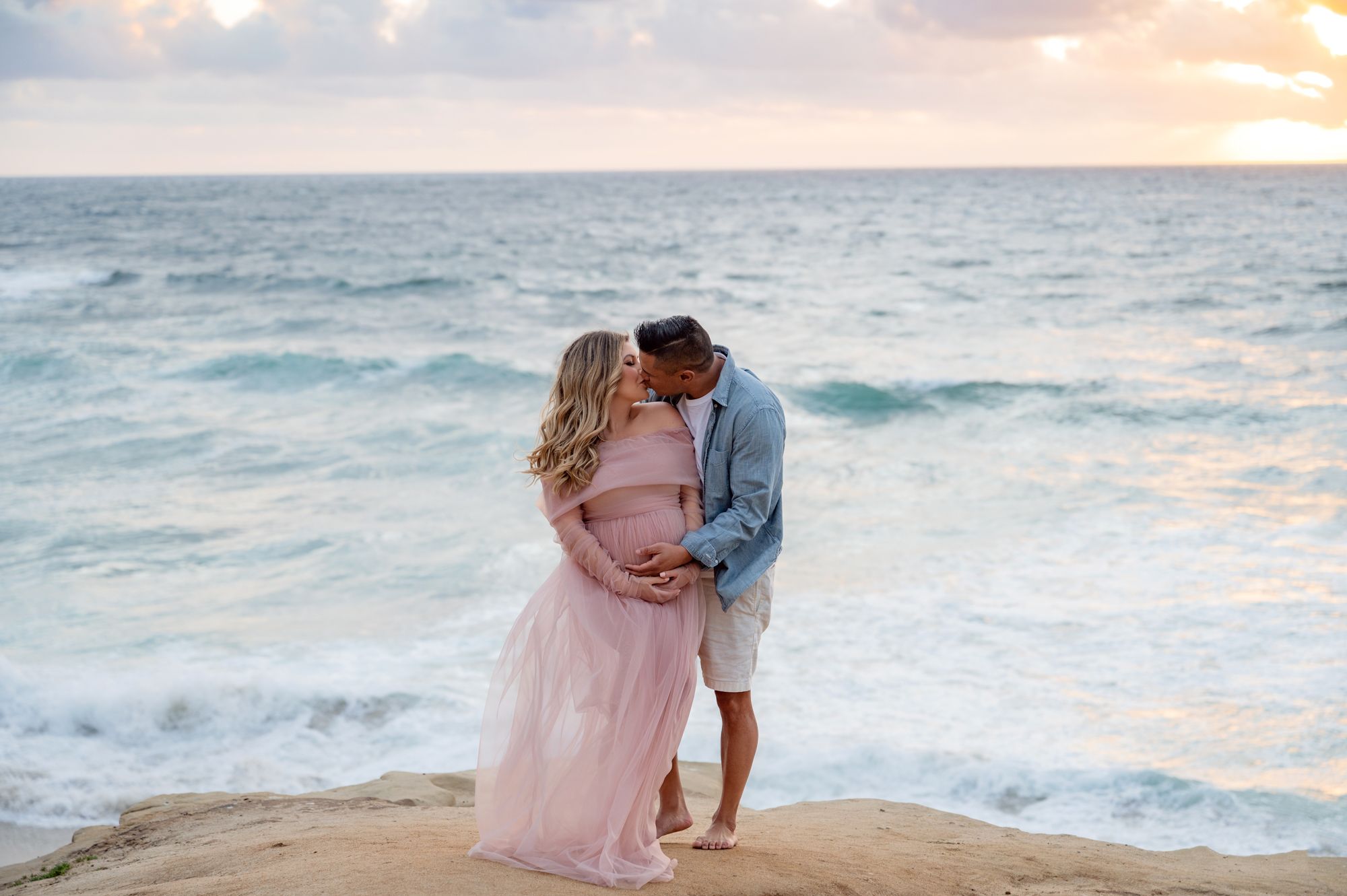 Couple sharing a kiss on coastal rocks at sunset, woman in pink maternity gown.