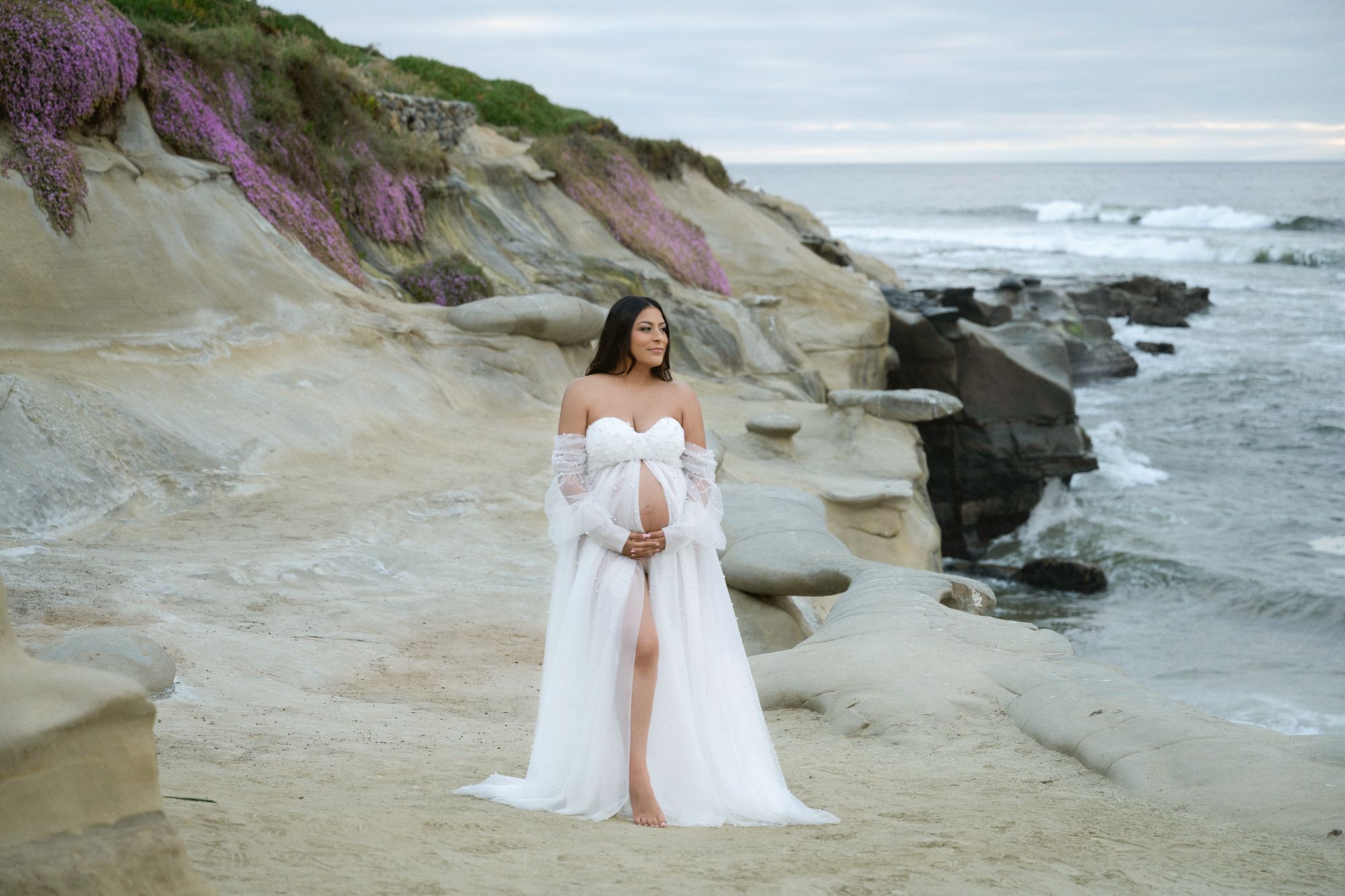 Pregnant woman in white off-shoulder gown posing on sandy beach with purple wildflowers.