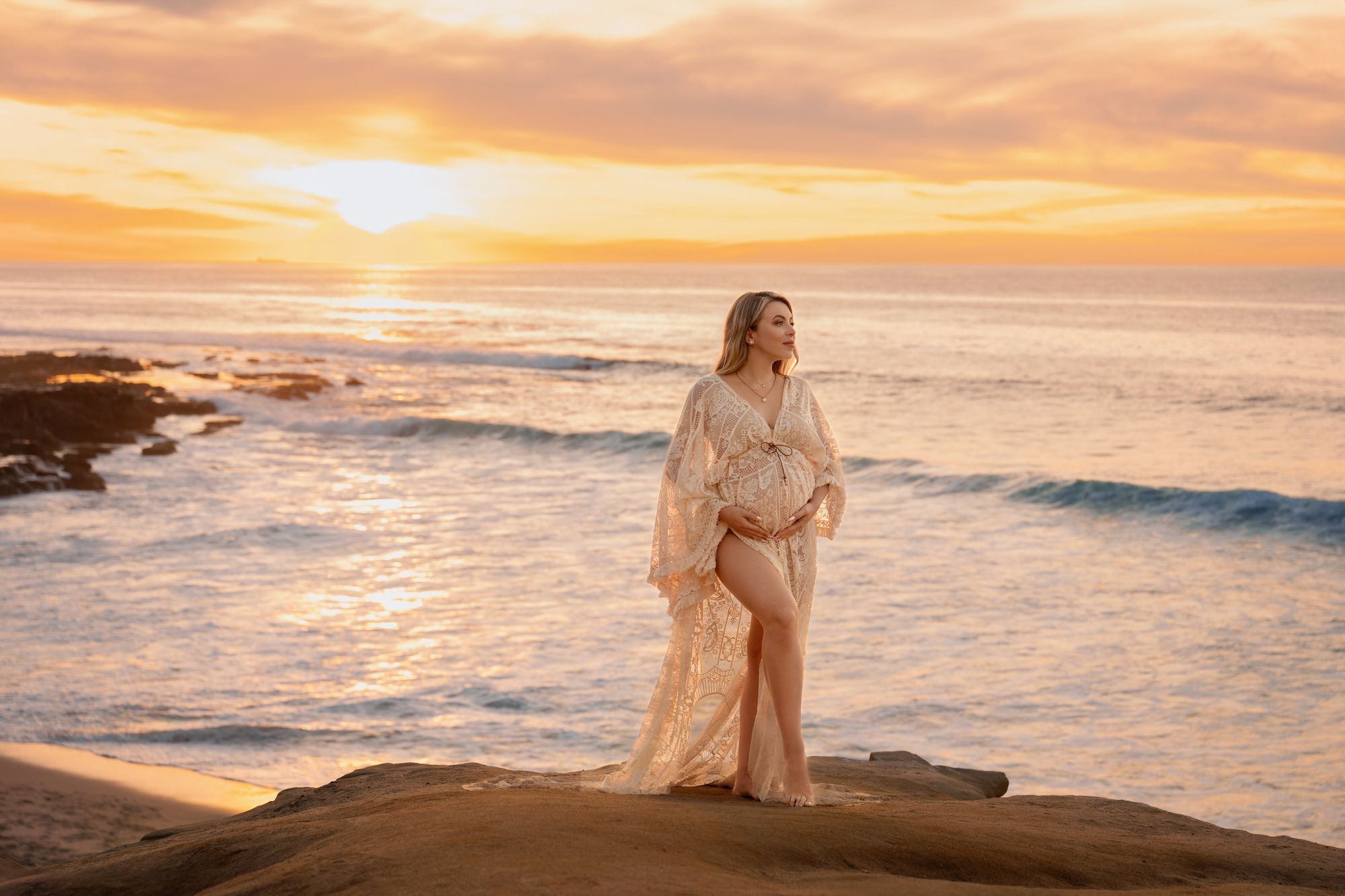 Maternity photo of woman in lace gown on rocky shore during vibrant orange sunset.