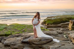 Pregnant woman in white flowing gown standing on mossy rocks at ocean sunset.