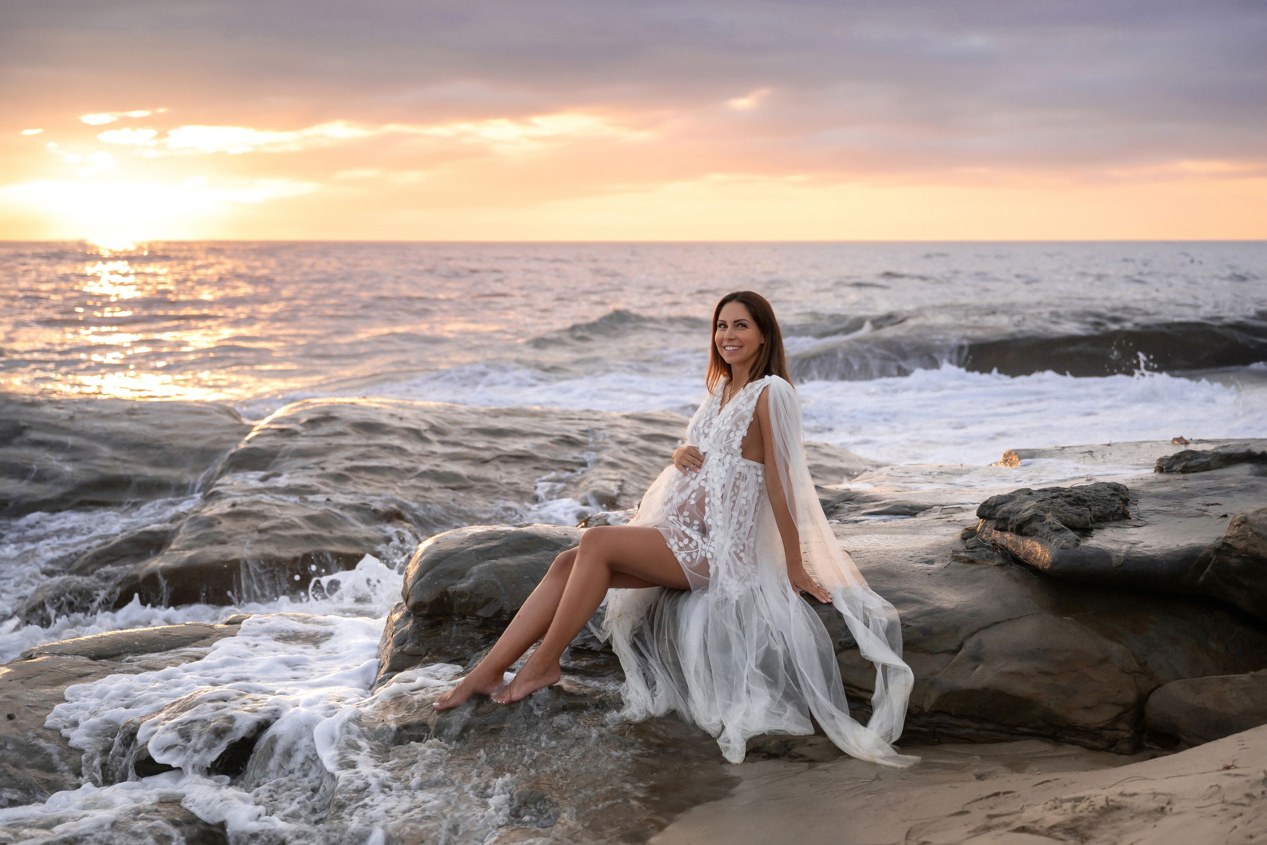 Pregnant woman in white lace gown sitting on coastal rocks during vibrant ocean sunset.