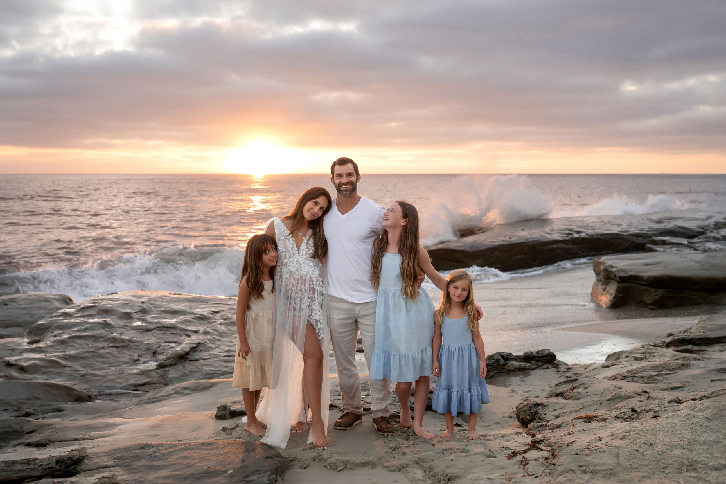 Happy family of five posing together on rocky beach at golden sunset by ocean waves.