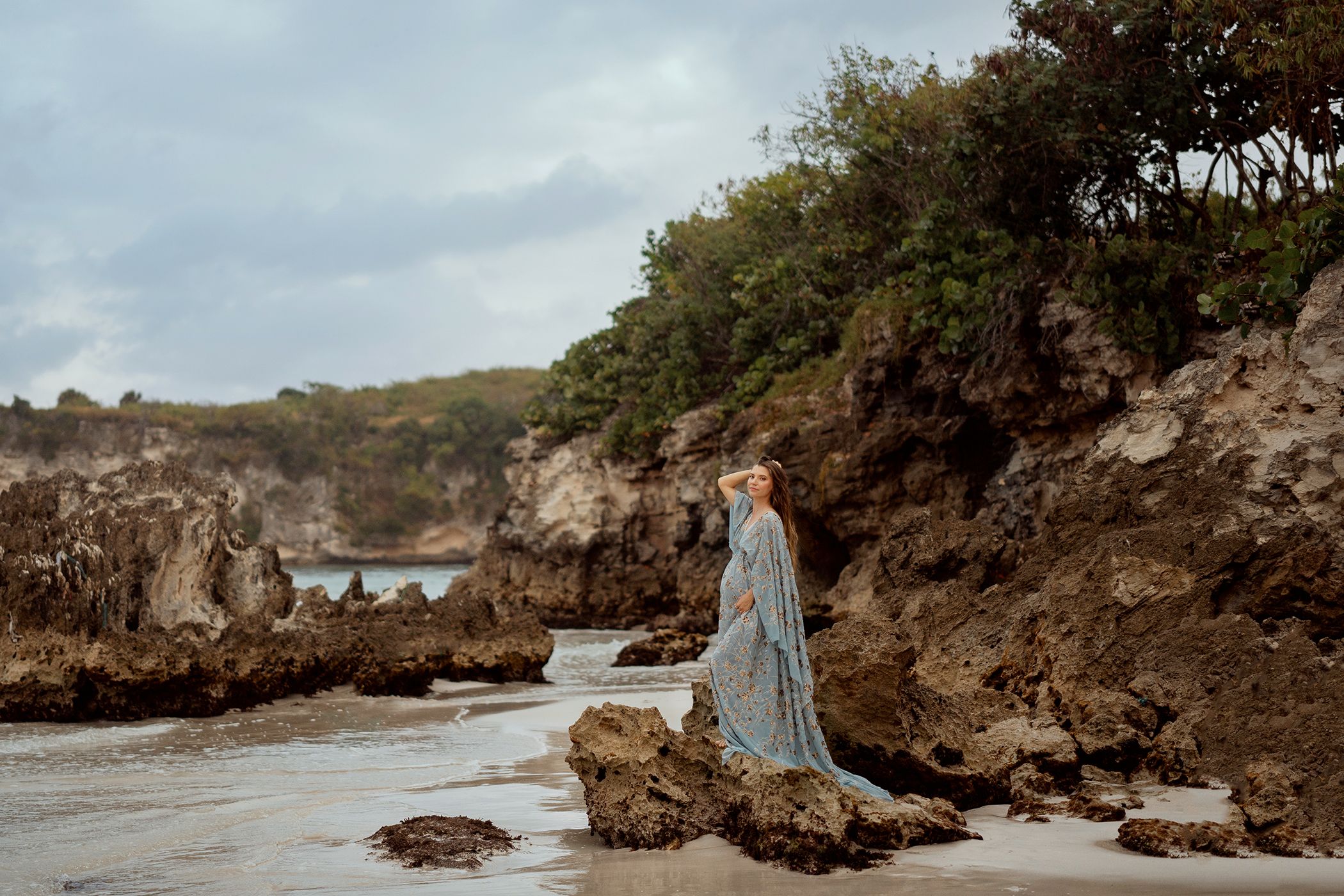 Woman in flowing blue gown posing on rocky beach with dramatic cliffs and cloudy sky.