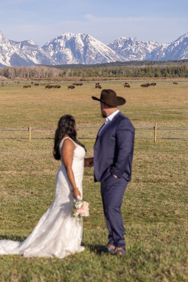 A couple in wedding attire poses in a field with snow-capped Grand Teton mountains creating a dramatic backdrop.