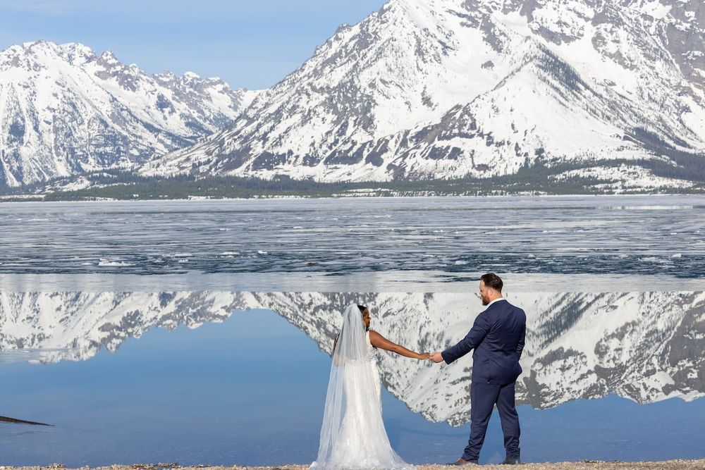 Couple in winter wedding attire poses together against snowy mountain peaks and frozen lake landscape.
