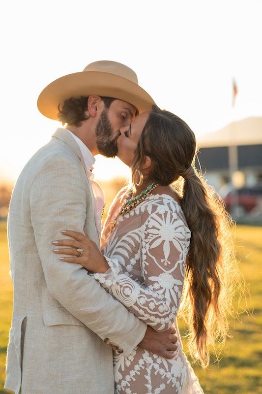 A couple embraces in a golden sunset field while wearing western and lace attire for their wedding photos.