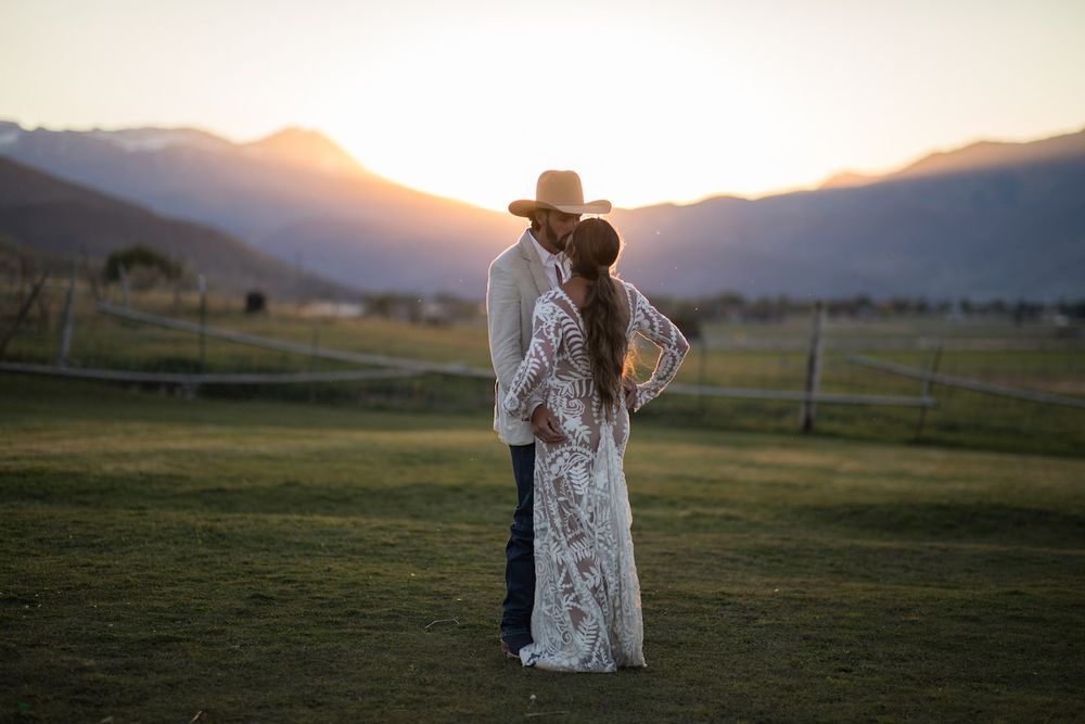 A couple embraces in a lace dress and suit against a golden sunset in a grassy field with mountains in the distance.