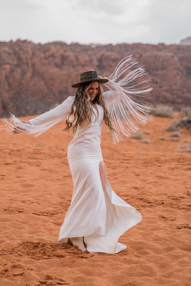 A figure in a flowing white dress and hat twirls gracefully against a backdrop of red desert rocks and sand.