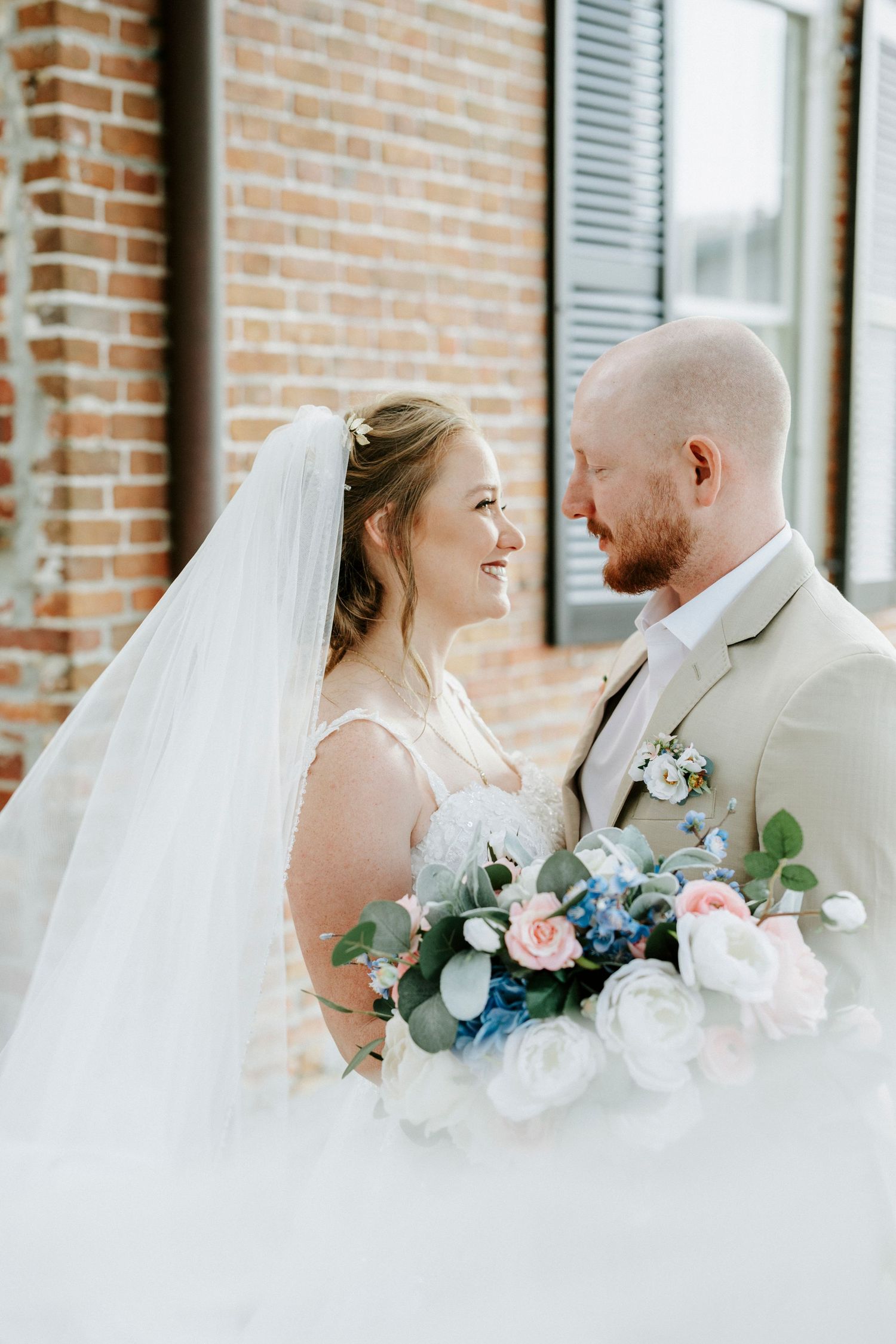 A wedding couple shares tender moments in front of a brick wall, holding pastel pink and blue floral bouquets.
