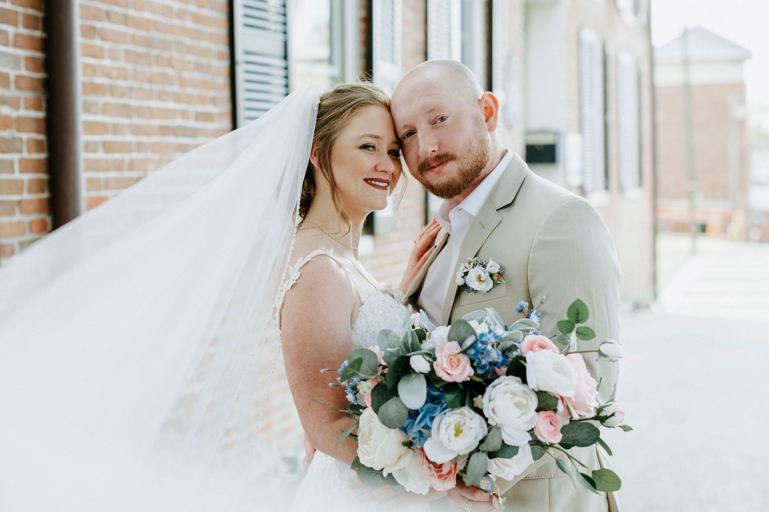 Romantic wedding couple embraces outdoors against brick wall with soft floral bouquet and natural lighting.