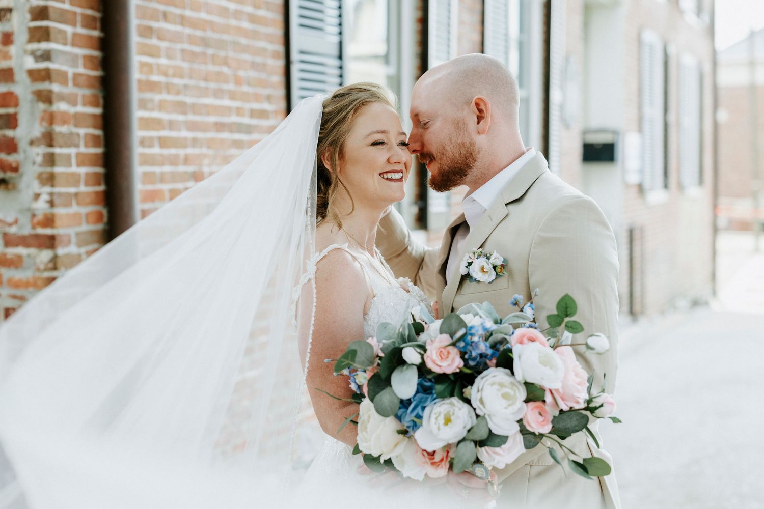 A romantic wedding moment captured against a brick wall backdrop with soft floral bouquet details.
