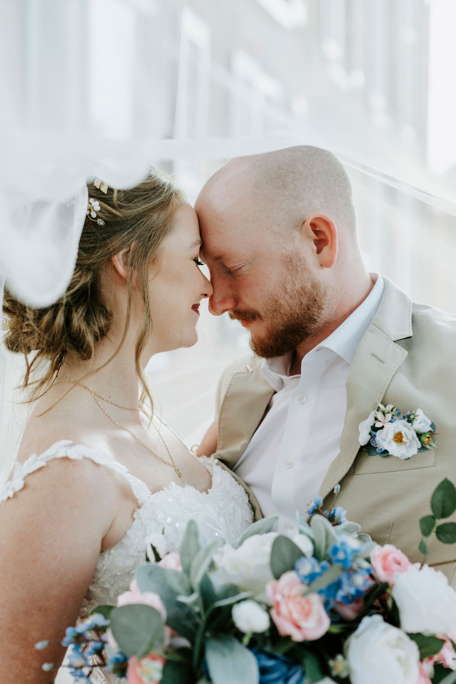 A romantic wedding couple shares an intimate moment with their foreheads touching while holding a pastel floral bouquet.