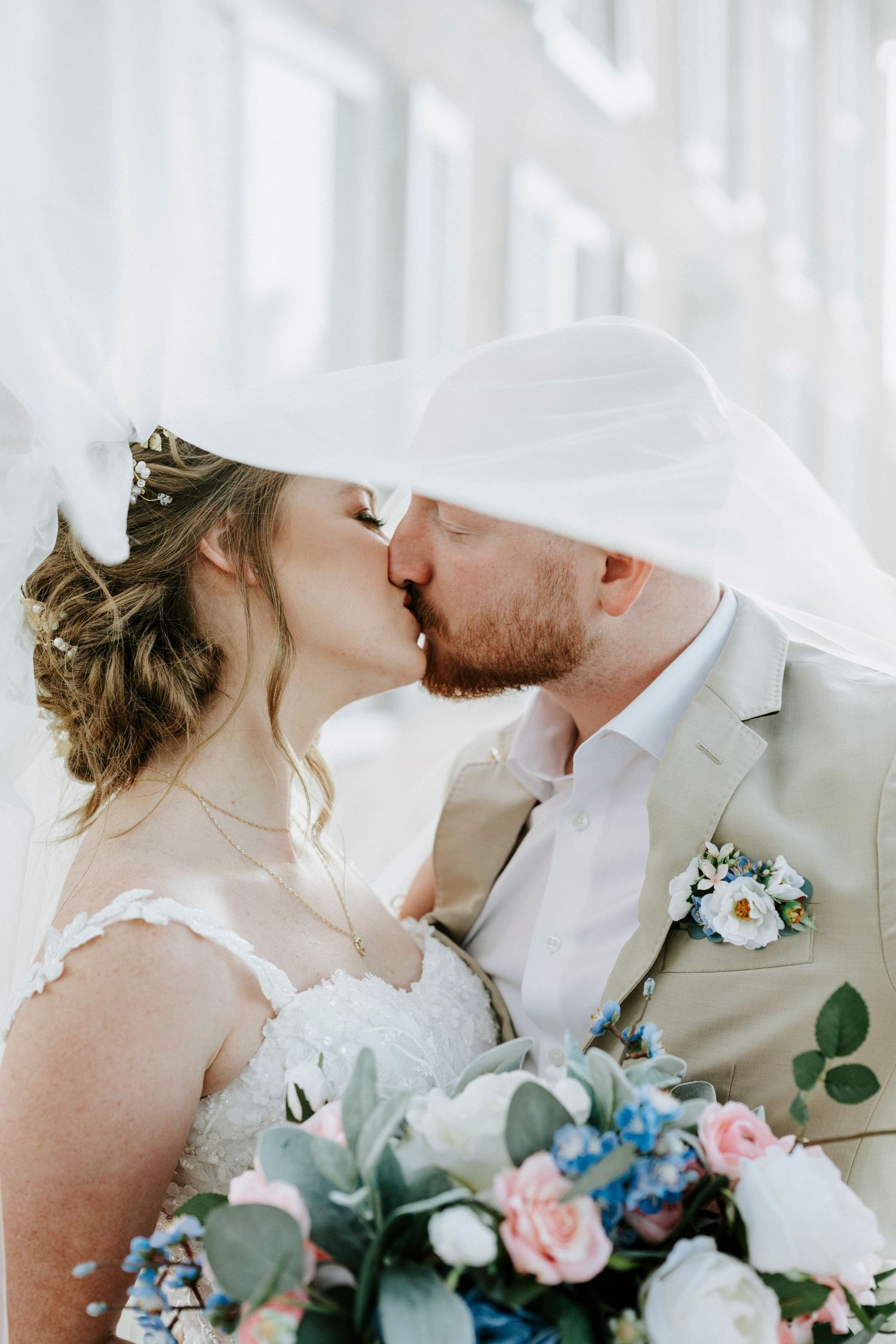 A romantic wedding couple sharing a tender kiss against a bright window backdrop with pink and blue floral bouquet.