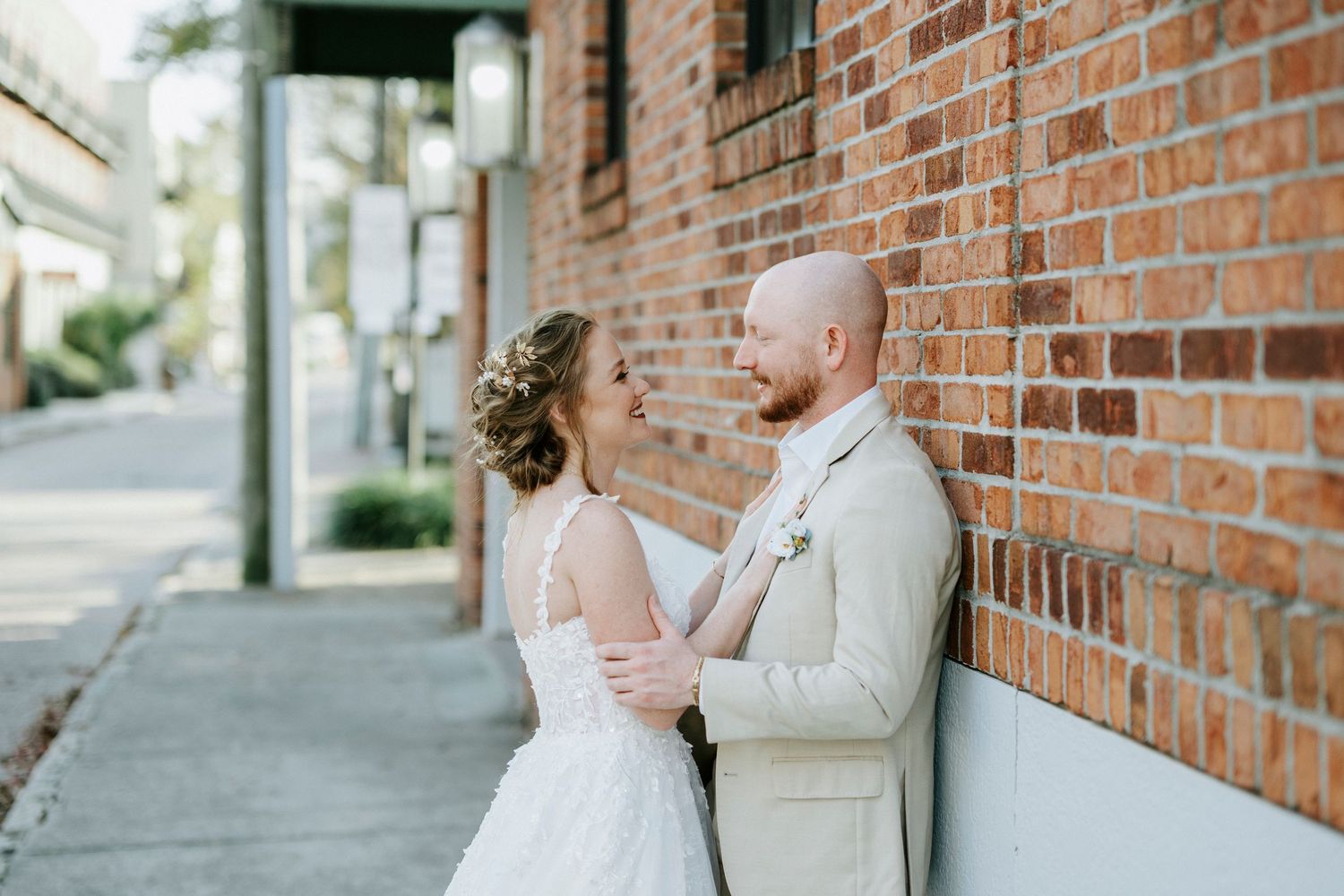 A romantic wedding couple embraces against a rustic brick wall on a sunny sidewalk.