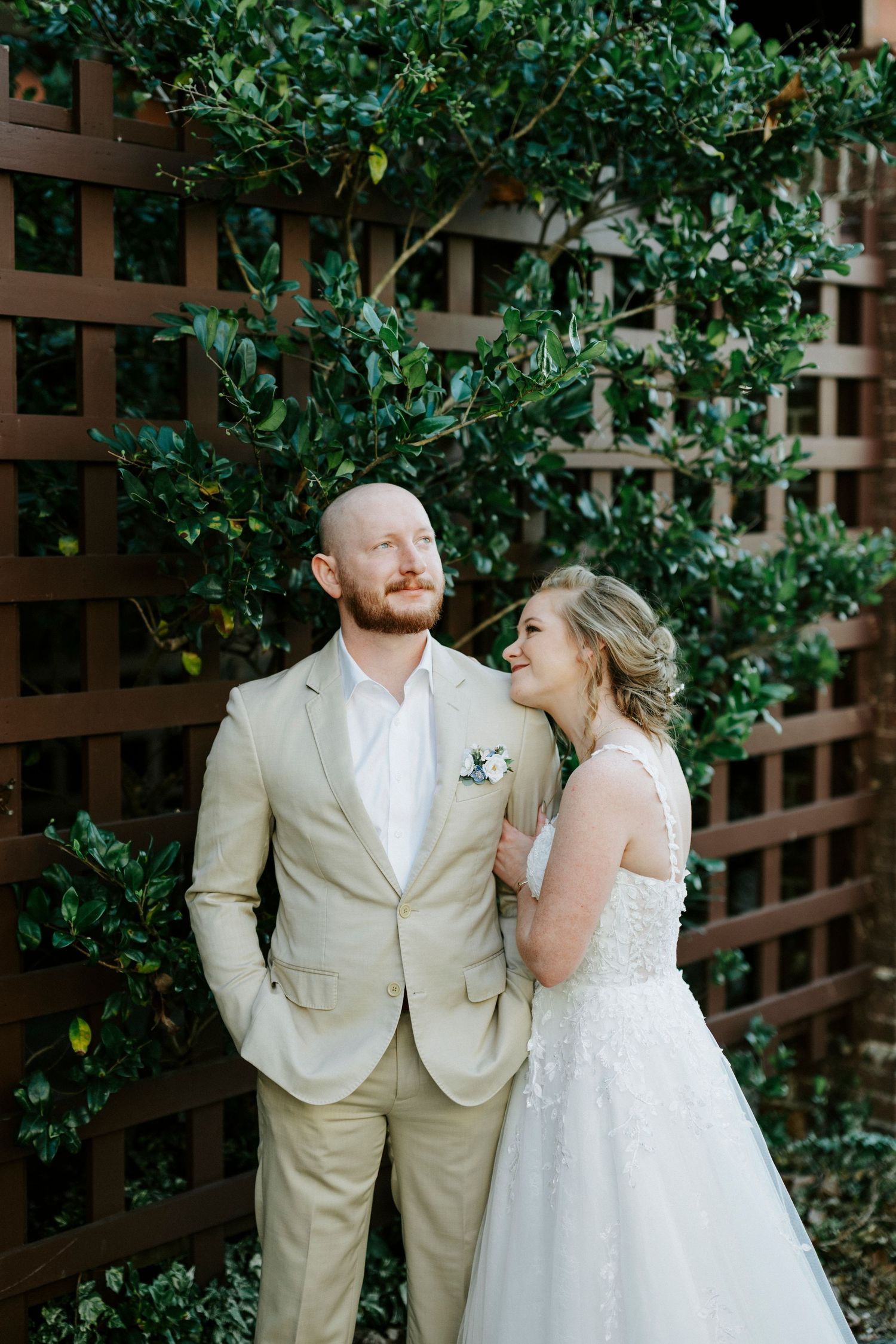 A wedding couple poses together against a wooden lattice backdrop with lush greenery.