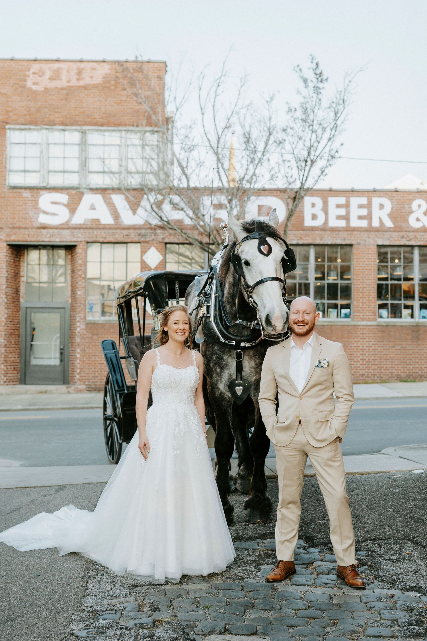 A wedding couple poses with a black horse in front of a brick industrial building with vintage signage.