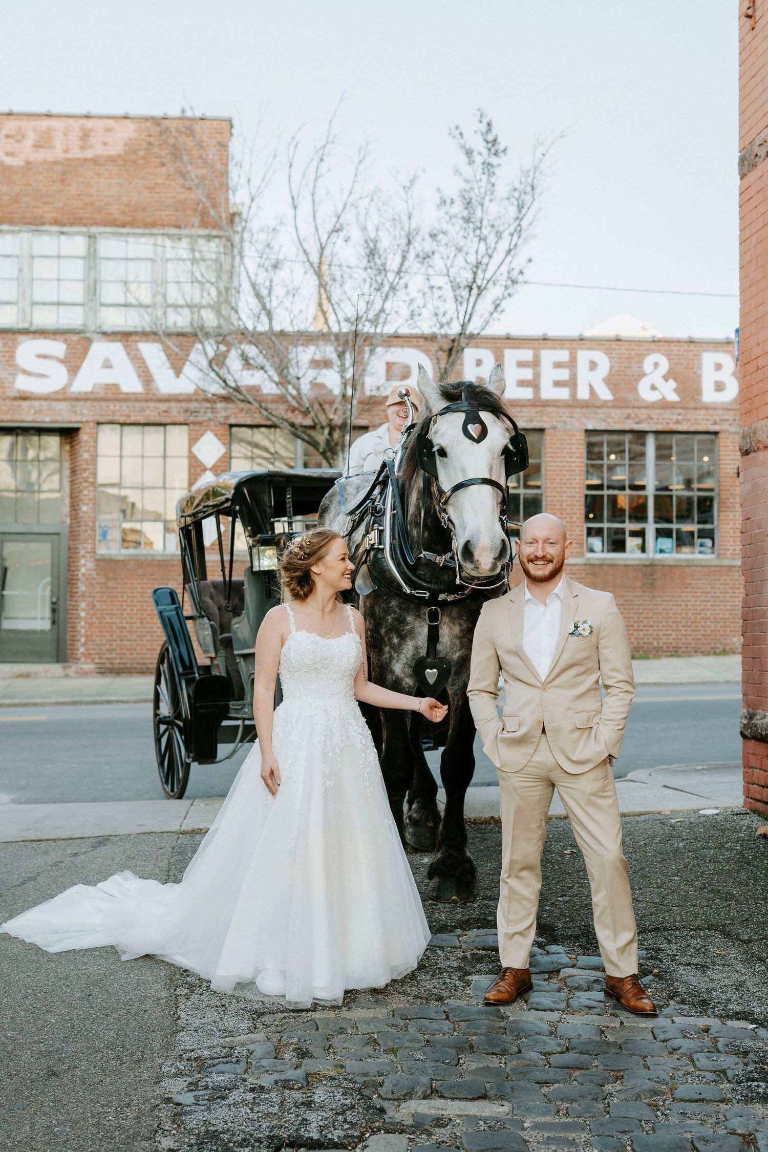 A wedding couple poses in front of a vintage brick building with a horse-drawn carriage.