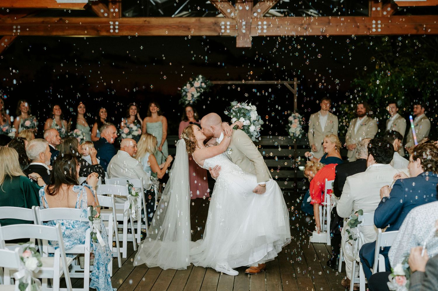 Wedding guests dance and celebrate during an evening reception under twinkling lights at a rustic venue.