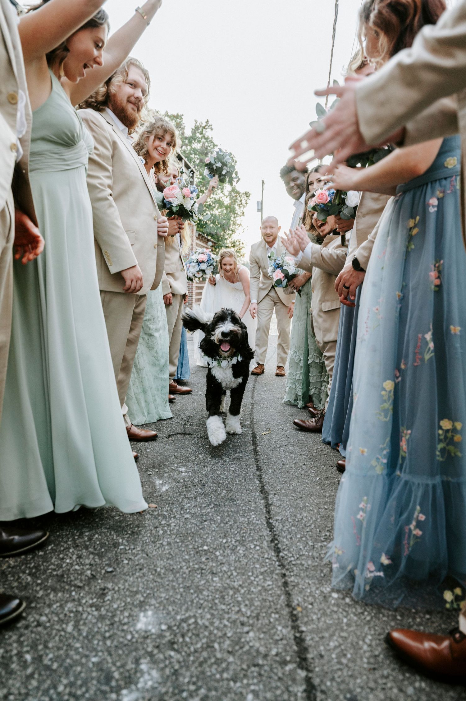 Wedding guests in blue and mint dresses stand on a gravel path during an outdoor celebration.