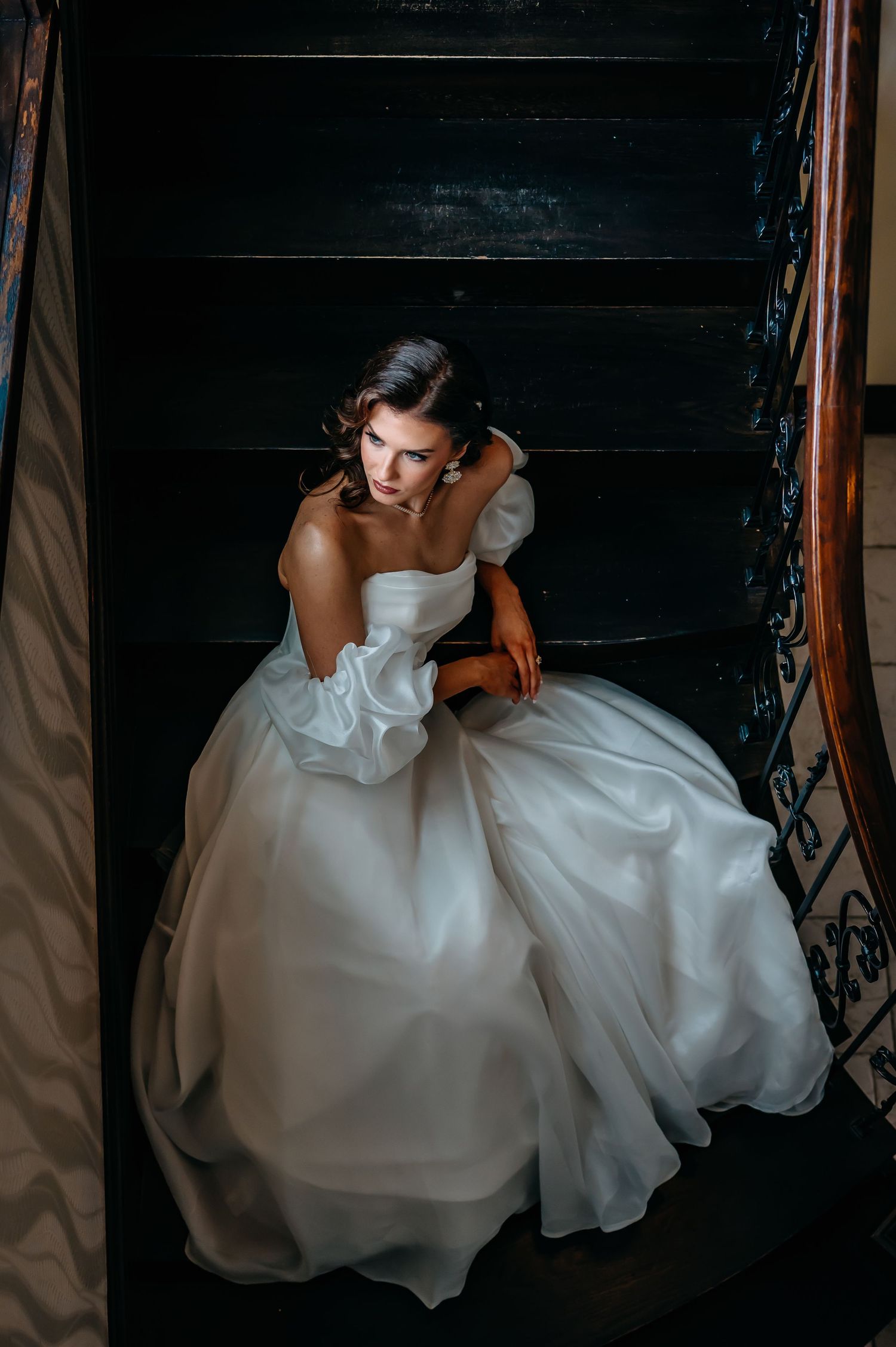 A bride in a flowing white gown sits gracefully on dark wooden stairs in dramatic lighting.