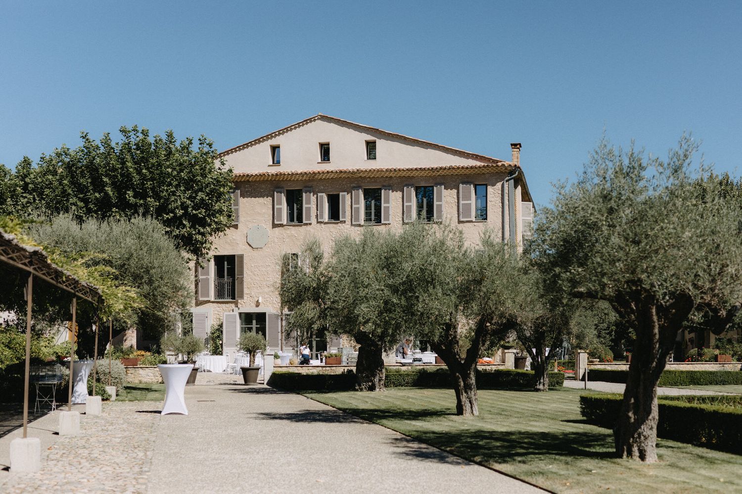 Elegant Italian villa with stone facade surrounded by olive trees and manicured grounds on a sunny day.