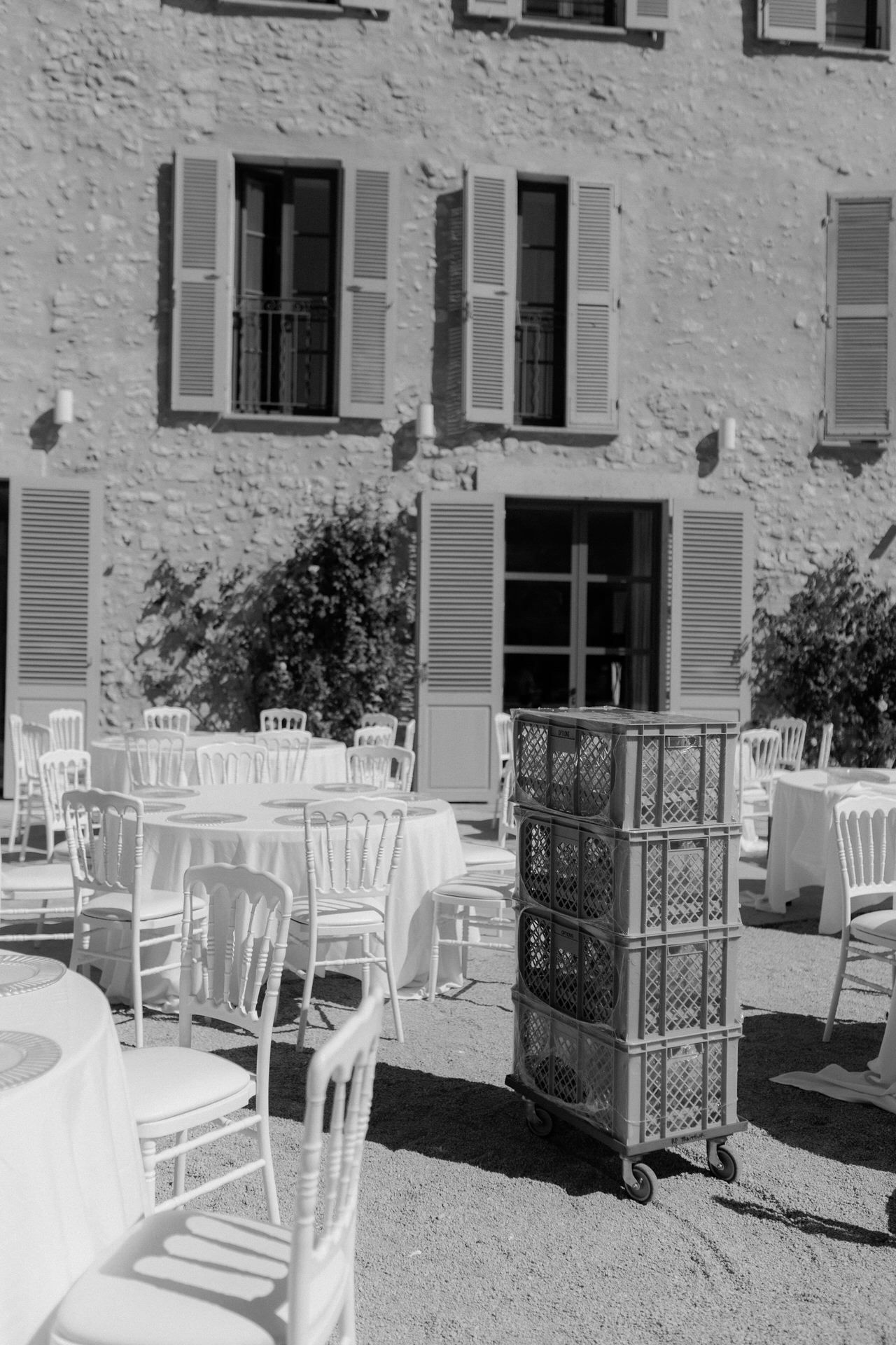 Outdoor cafe seating with white chairs and tables arranged in front of a Mediterranean-style building with shuttered windows.