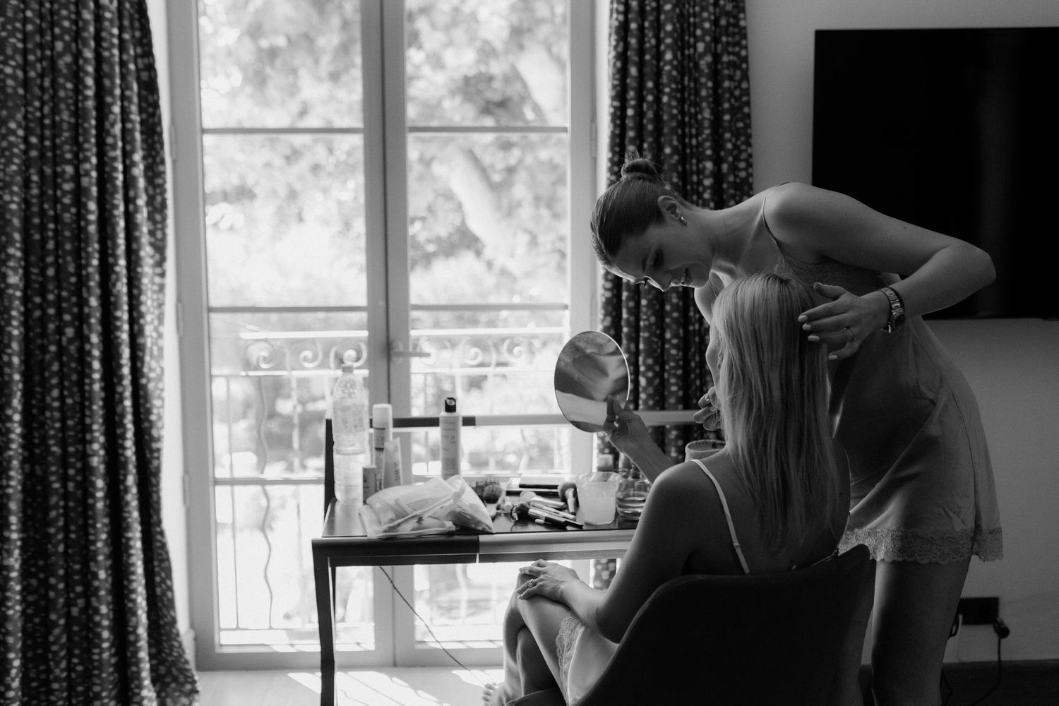 A black and white photo showing a tender moment between two people in front of a window with curtains.