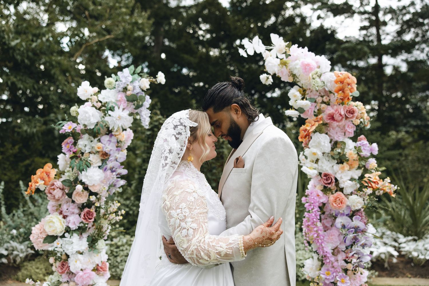 A couple shares a romantic moment under a beautiful floral arch decorated with pink and white flowers in an outdoor garden setting.
