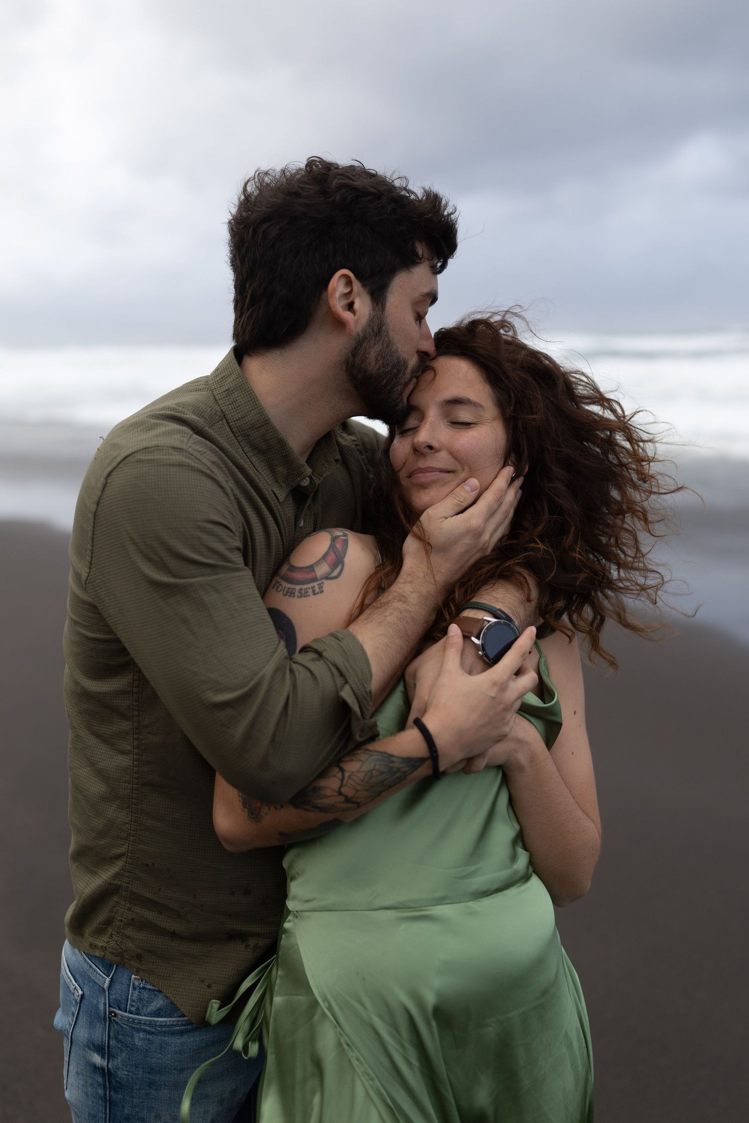 A couple embraces passionately on a cloudy beach, with the woman wearing a green dress and the man in olive shirt and jeans.