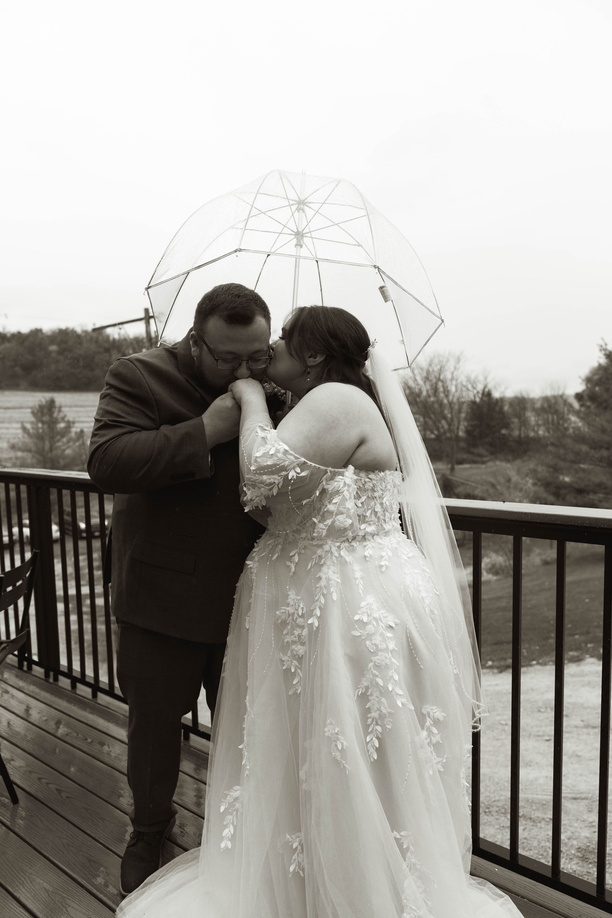 A romantic moment shared under a clear umbrella on a deck with a scenic backdrop.