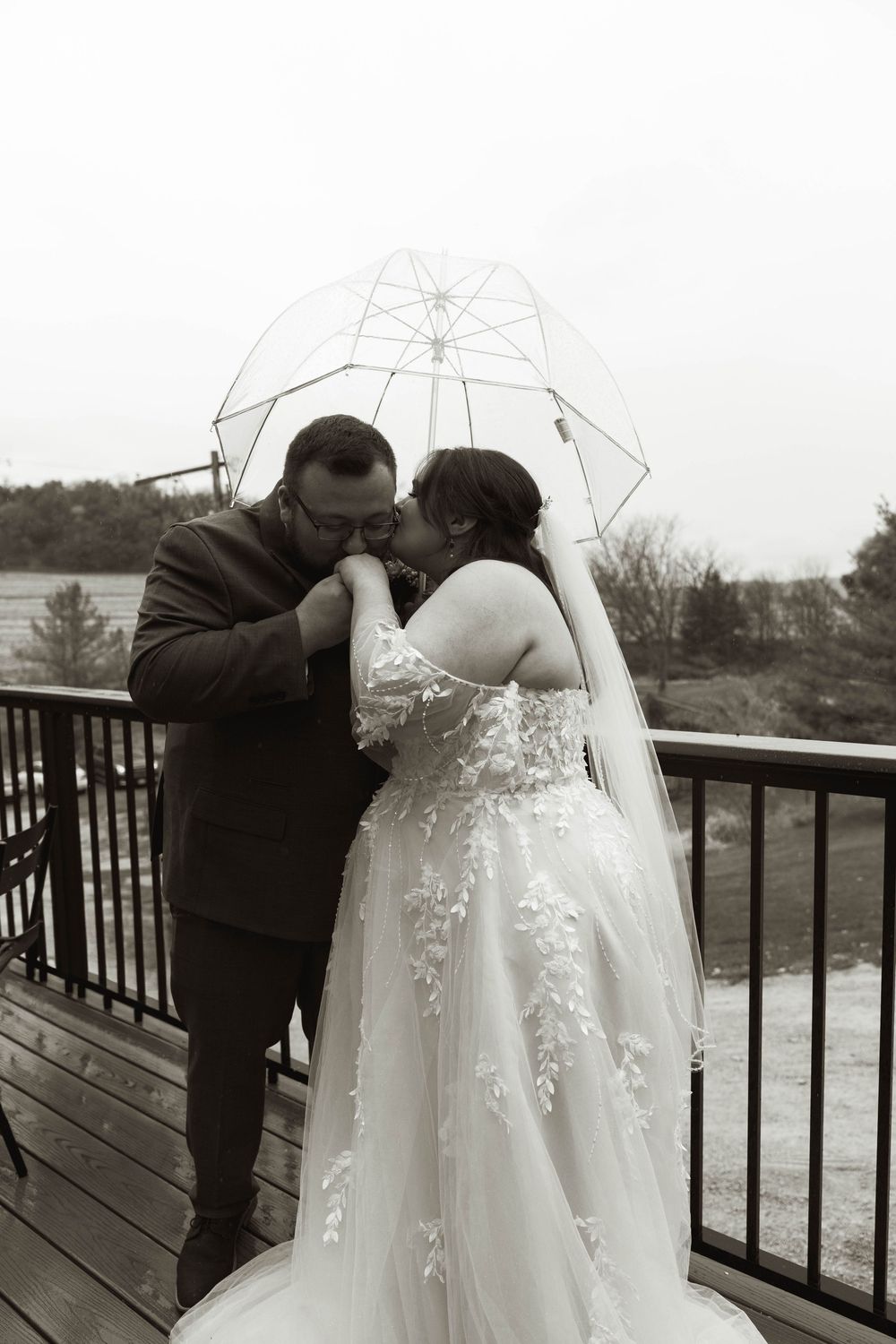 A romantic moment shared under a clear umbrella on a deck with a scenic backdrop.