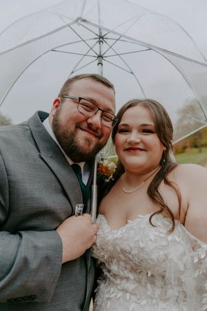 A cheerful wedding portrait of newlyweds smiling together under a clear umbrella outdoors.