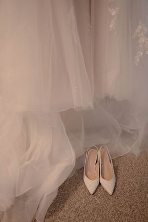 White wedding shoes placed beneath flowing tulle fabric on beige carpet.