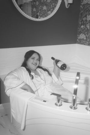 A person relaxes in a bathtub holding a wine bottle while smiling in a black and white photograph.
