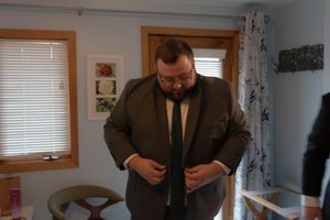 A groom adjusts his tie while getting ready in a room with wooden window frames.