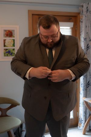 A person in a dark suit adjusts their clothing while standing in a room with floral curtains.