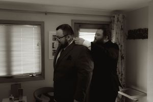 Two people in formal attire stand in a room with venetian blinds and curtains.