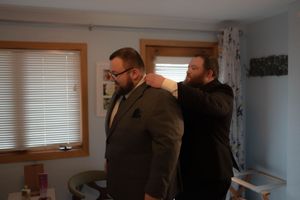 Someone helps adjust a suit jacket in a room with natural lighting and floral curtains.