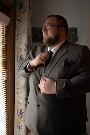 A person in a grey suit and glasses adjusts their tie while standing by a window.