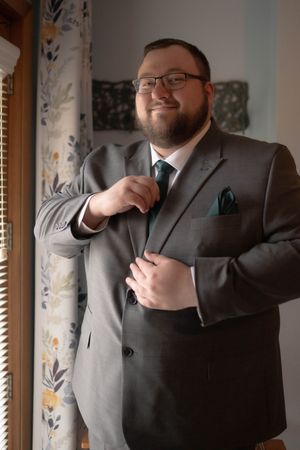 A person in a grey suit and blue tie smiles while adjusting their formal attire.