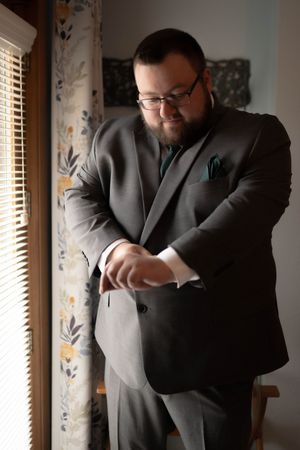 A person in formal wear checks their watch while standing by a window with floral curtains.
