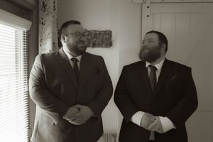 Two people in dark suits stand together in a room with natural lighting.