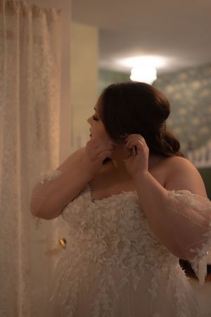 A bride gets help putting on her wedding dress in a softly lit room.
