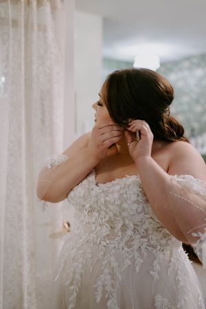 A bride in a lace wedding dress puts on earrings while getting ready.