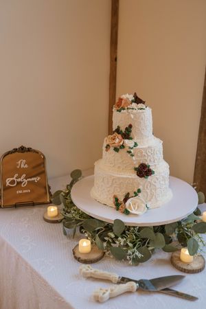 White tiered wedding cake decorated with roses and greenery on display with rustic decor.