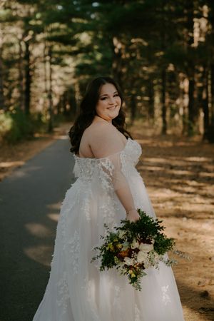 Bride in lace wedding gown holds bouquet on forest path in golden light.